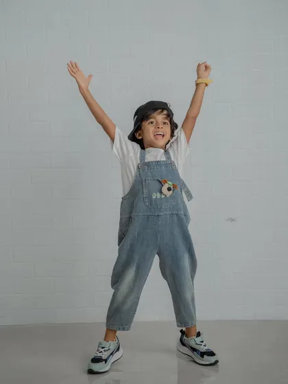 An energetic shot of a boy in overalls and a cap with his arms raised in excitement against a white brick wall.