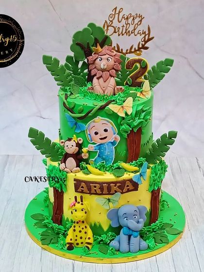 A wild two-tier cake combining a jungle theme with Cocomelon for a 2nd birthday. It features JJ alongside a fondant lion, monkey, giraffe, and elephant amidst lush greenery.