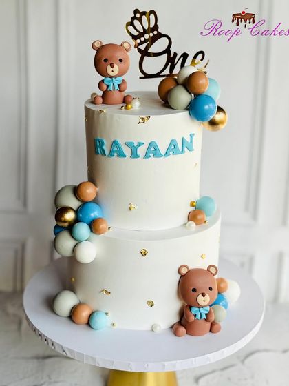 A charming two-tier teddy bear cake for Rayaan's first birthday. The clean white cake is decorated with blue, brown, and white balloons and two adorable fondant bears.