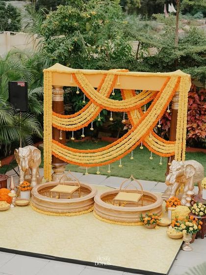 A wider view of the marigold-themed Haldi stage. The setup is framed by lush greenery, making the bright yellows and oranges of the flowers pop. The golden elephants add a touch of regal charm.
