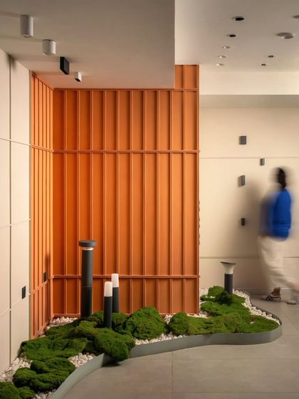 The motion-blurred figure gives a sense of movement and scale to the space. The design combines natural elements like moss with modern industrial materials and bold orange fluted panels.