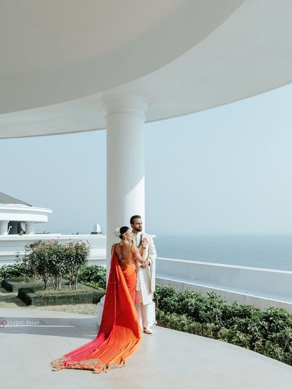 A breathtaking shot of the couple in Vietnam, with the bride's vibrant orange and red saree flowing behind her against the backdrop of the sea and a white villa.