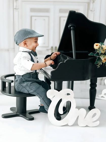 A dapper little gentleman playing the piano for his first birthday portrait. The classic outfit with suspenders and a cap, paired with the elegant white studio backdrop, creates a timeless photo.