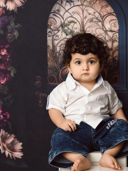 A handsome little boy with curly hair sits for his portrait against a beautiful floral backdrop.