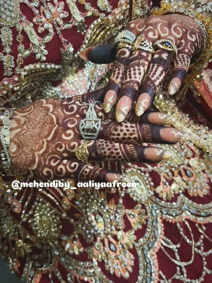 A close-up of the bride's hands, showcasing the detailed finger work and rich henna stain against her ornate dress.