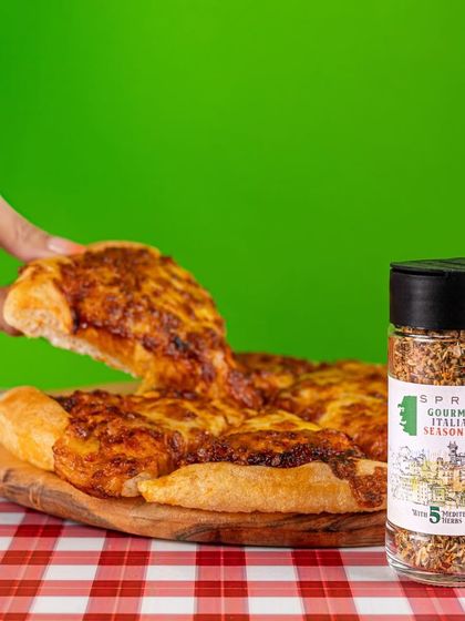 A vibrant and crisp shot of Italian seasoning next to a cheesy pizza. The bright green background provides a strong color block effect, ensuring the product is the center of attention while still being in a relevant context.