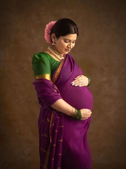A beautiful and introspective portrait of a mother-to-be in a traditional purple saree, her hands gently cradling her bump. The soft lighting creates a painterly, classic feel.