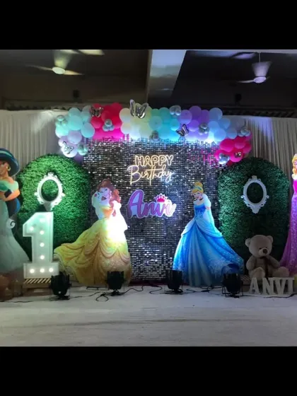A Disney princess themed first birthday stage, with cutouts of various princesses, a green hedge backdrop, and a light-up number '1'.