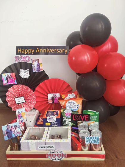 This anniversary hamper uses a striking red and black theme to create a romantic mood. It's filled with snacks, drinks, and decorated with personal photos and paper fans, perfect for a cozy celebration.
