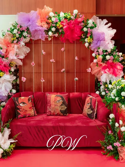 A vibrant seating area for a Mehendi, with a red sofa and cushions featuring traditional Indian art, set against a colorful floral arch.