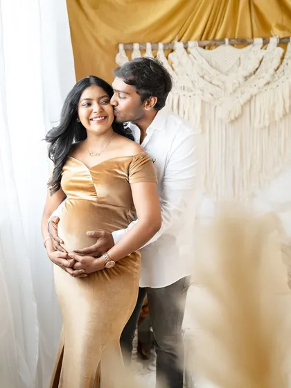 A sweet and affectionate moment captured in a bright, boho-style room. The partner embraces the mom-to-be from behind, kissing her cheek as she smiles, both celebrating the baby on the way.
