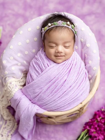 A smiling newborn in a lavender wrap, looking angelic in a basket surrounded by purple flowers.