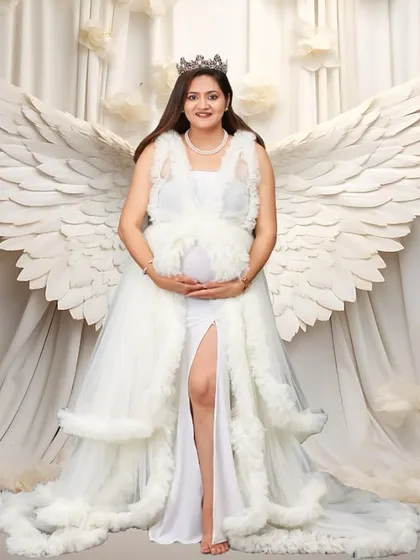 A powerful and angelic studio portrait of an expectant mother wearing a beautiful white gown and a crown. She stands gracefully with large, white angel wings behind her against a dreamy, cloud-like backdrop, portraying the miracle of motherhood.
