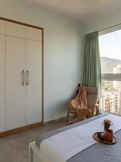 This bedroom with a large window and stunning city view is kept minimal and airy with a simple white and wood wardrobe and a comfortable rocking chair.