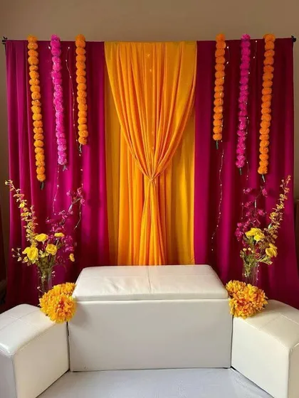 For an intimate at-home Haldi ceremony, we created this simple yet elegant backdrop. It uses vibrant pink and orange drapes with hanging marigold garlands for a traditional and festive feel.