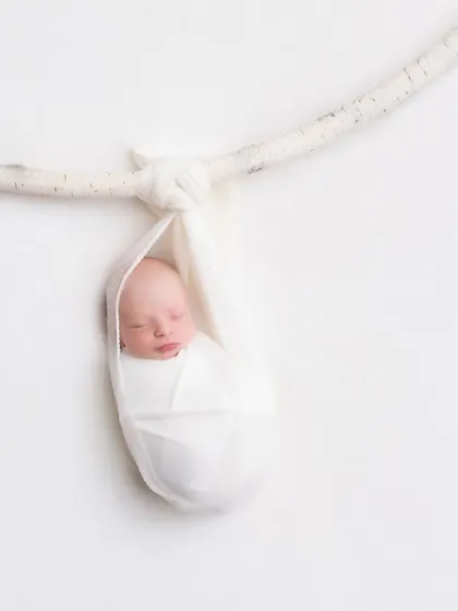A newborn baby, swaddled snugly in white, sleeps soundly in a hanging sling against a clean, minimalist background. This artistic shot highlights the delicate and trusting nature of a newborn, emphasizing safe and creative posing.