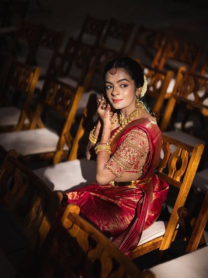 A bride seated in a dramatic pose, surrounded by empty chairs. Her red saree stands out, symbolizing her as the center of the celebration.