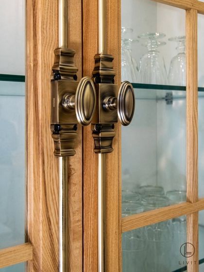 A detailed view of our Cremone Bolt on a glass-fronted kitchen cabinet. This classic hardware adds a layer of vintage charm and mechanical beauty, perfectly complementing the antique brass knobs and pulls used elsewhere in the space.