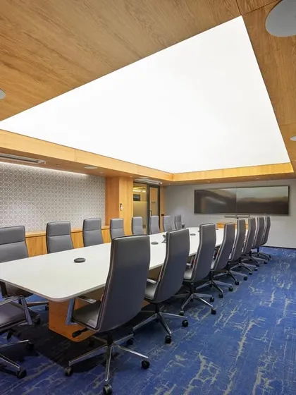 A large, formal boardroom featuring a stretch-lit ceiling that provides bright, even, and glare-free light. The room is equipped with a long conference table, comfortable seating, and an additional bench along the wall for larger meetings.