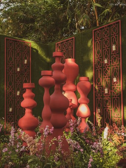 Red sculptural totems and jali screens are surrounded by flowers, creating artistic installations within the garden.