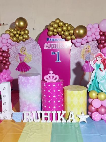 A full-stage princess-themed first birthday. The decor includes a rainbow-colored floor, princess cutouts, and a large light-up number '1'.