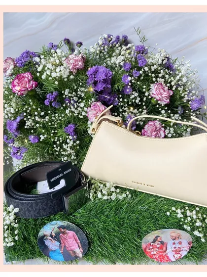 A luxurious anniversary gift featuring a Michael Kors belt and a Charles & Keith handbag, beautifully arranged on a bed of fresh flowers with personalized photo coasters of the couple.