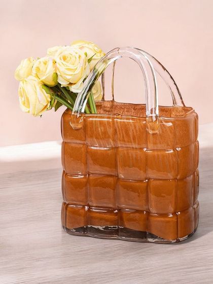 A unique vase handblown to resemble a woven handbag. The warm amber color and textured appearance make it a stylish and unconventional piece for holding flowers or as a decorative object.