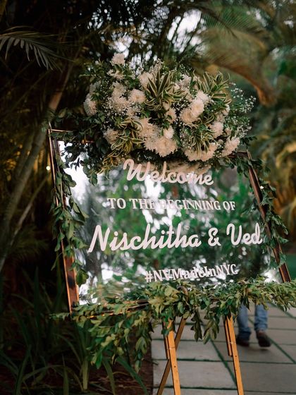 A personalized welcome to the Sangeet celebration. This mirrored sign, framed with lush greenery and white florals, adds a touch of modern elegance to the event entrance.