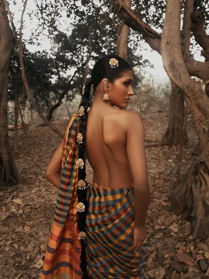 A beautiful back portrait from our 'Divine' series, shot in a natural forest setting. The focus is on the traditional checked saree and the intricate gold jewelry adorning the model's long braid.