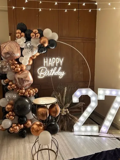 A modern and elegant setup for a 27th birthday at home, featuring a rose gold, black, and white color palette with a neon sign and marquee numbers.