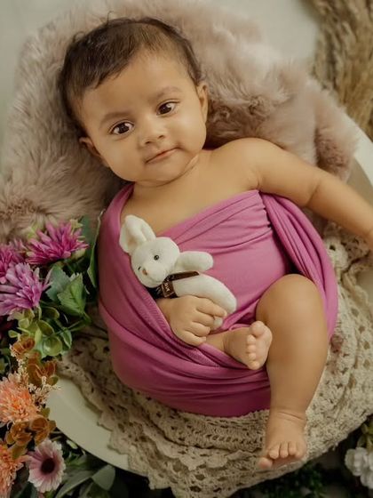 An awake and curious newborn, wrapped in a magenta cloth, holds a small bunny toy while nestled in a floral arrangement.