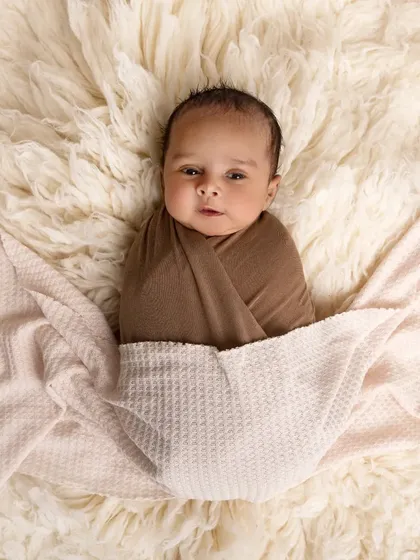 A newborn is wrapped in a brown swaddle and tucked under a waffle-knit blanket. The soft, neutral tones and textures create a feeling of warmth and comfort.