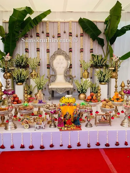 An elegant and bright Seemantha decoration featuring a white backdrop with hanging sugandharaja garlands. The stage is flanked by banana plants and a symmetrical arrangement of offerings, creating a pure and sacred ambiance.
