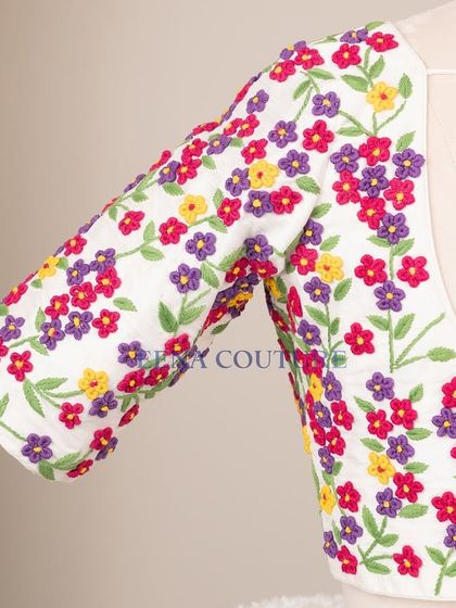 A garden of flowers on a blouse. This white raw silk piece is covered in vibrant, multi-colored floral French knot embroidery, making it a cheerful and eye-catching addition to any festive wardrobe.