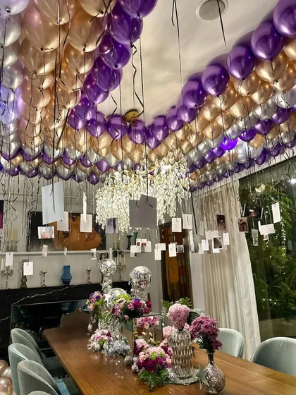 A glamorous and personal birthday setup for Janhvi Kapoor. The entire ceiling is covered in metallic purple and gold balloons, with personal photos hanging down over an elegantly styled dining table, creating a room filled with memories.