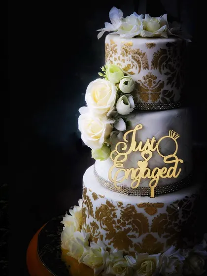 A luxurious three-tiered engagement cake featuring an intricate gold damask stencil pattern and elegant white sugar roses.