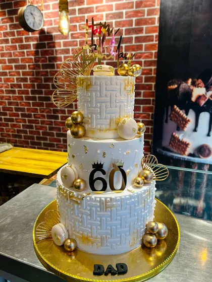 Celebrating a 60th birthday in style with this grand three-tier cake. The design features a white basket-weave texture, gold spheres, macarons, and elegant fan details for a truly luxurious feel.