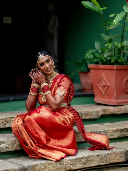 A bride in a radiant red saree, sitting on stone steps, adjusts her jewelry in a moment of quiet preparation.