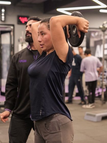 A trainer assists a member with an overhead tricep extension using a weight plate, ensuring correct form to isolate the tricep muscles.
