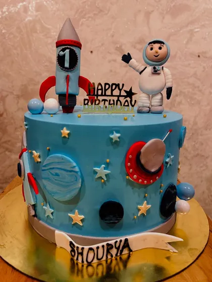 A space-themed cake for Shourya's first birthday, featuring a fondant astronaut, rocket, and planets.