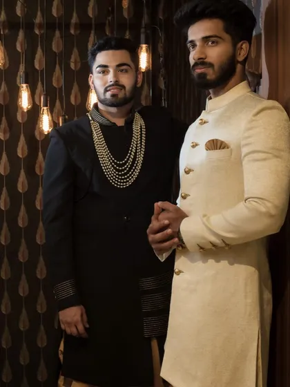 A groom and his best man, showcasing two different but complementary styles. One in a classic off-white bandhgala and the other in a black sherwani with a statement pearl necklace.