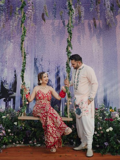 A fairytale-like shot of the couple at their Mehendi. The bride is on a floral swing while the groom stands beside her, creating a whimsical and romantic scene.