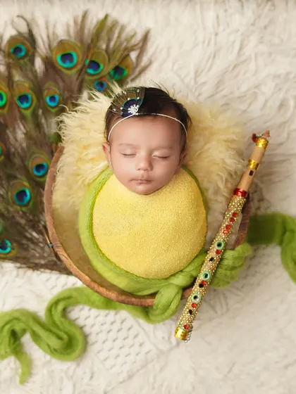 The full 'Baby Krishna' setup, with the newborn in a basket, surrounded by peacock feathers and holding a flute (bansuri).