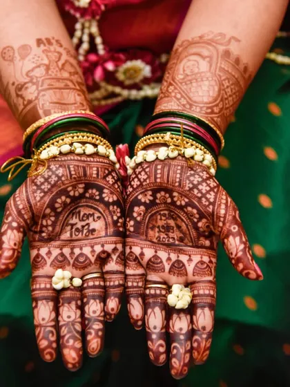 The hands of a beautiful mom-to-be, showcasing her intricate baby shower mehendi. The design includes 'Mom To Be' and the couple's anniversary date.