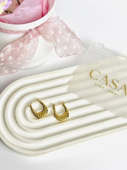 A pair of textured gold hoop earrings styled on a modern tray. The simple composition and bright lighting make the product details pop.