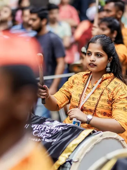 A female dhol player with a focused expression, her stick poised to strike the drum, captured in the midst of the procession.