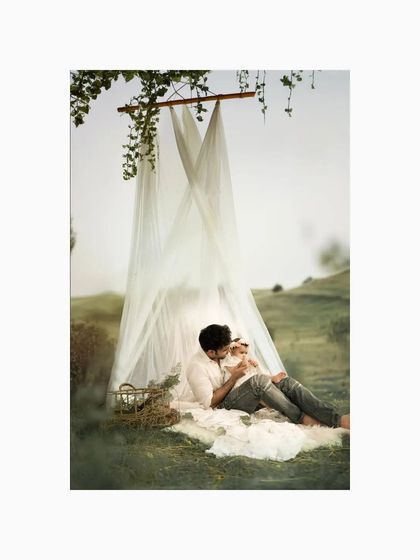 A father lies on a soft blanket outdoors, cradling his baby. The dreamy, natural setting with flowing drapes creates a peaceful and artistic scene for this first birthday photoshoot.