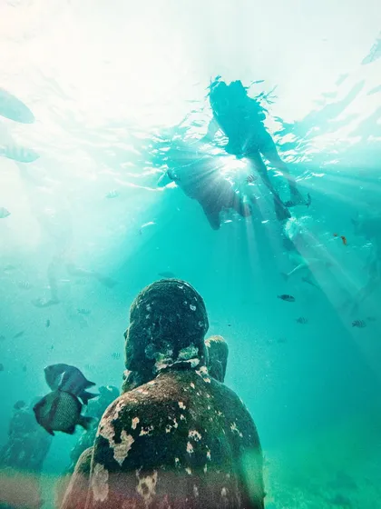 A surreal underwater shot of a swimmer seen from below, with sun rays piercing the water near the Gili Meno statues in Bali.