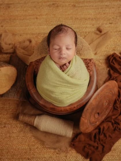 A newborn wrapped in light green sleeps in a wooden bowl, part of a rustic flat-lay composition with wood and brown fabrics.