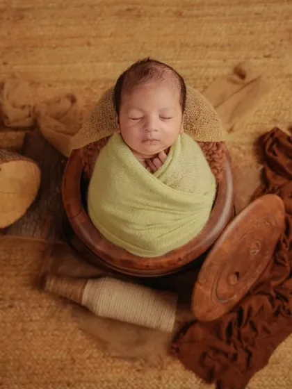 A newborn wrapped in light green sleeps in a wooden bowl, part of a rustic flat-lay composition with wood and brown fabrics.